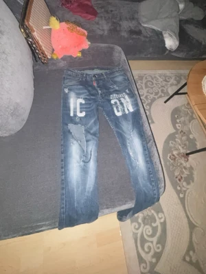 Blå Dsquared2 ICON jeans - Säljer ett par blå jeans från Dsquared2 med stor vit 'ICON'-text på framsidan. Jeansen har slitna detaljer, raka ben och klassisk femficksmodell. Materialet är denim i bomull och de har en cool streetwear-vibe.