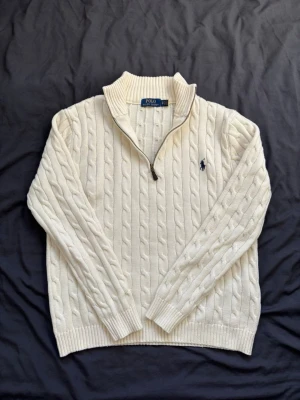 Vit kabelstickad tröja Polo Ralph Lauren - Kabelstickad vit tröja från Polo Ralph Lauren med halv dragkedja och hög krage. Tröjan har det klassiska mörkblå logobroderiet på bröstet och ribbade muddar vid ärmslut och nederkant. Perfekt för lager på lager och snygg till jeans.