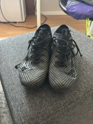 Nike svartgrå fotbollsskor - Säljer ett par snygga fotbollsskor från Nike i svart och grått med ett mönster av tunna ränder. Skorna har snörning och låg profil, samt en tydlig Nike-logga på sidan. Perfekta för dig som vill ha stilrena och funktionella skor på planen. 