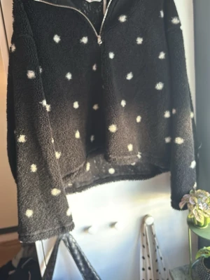 Svart prickig teddytröja med dragkedja - Mysig svart teddytröja med vita prickar och halv dragkedja framtill. Tröjan har lång ärm och är gjord i fluffigt material som håller dig varm. 