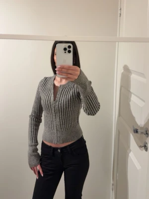 Grå ribbad kofta med dragkedja - Säljer en gråmelerad ribbstickad kofta med dragkedja framtill. Modellen är croppad med långa ärmar och bred mudd i midjan och vid ärmsluten. Perfekt för lager på lager och enkel att matcha med olika outfits.