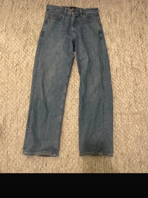 Blå raka jeans från Lee - Klassiska blå jeans från Lee med rak passform och hög midja. Jeansen har fem fickor, bälteshällor och Lee-detaljer på knapp och etikett. Tillverkade i slitstark denim, perfekta för en avslappnad och tidlös stil.
