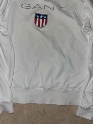 Vit sweatshirt från GANT - Vit sweatshirt från GANT med broderad logga och ett stort emblem med stjärnor och ränder på bröstet. Tröjan har rund halsringning och långa ärmar. Klassisk och stilren design som passar till mycket.Storlek S/M . Tröjan har ett litet litet hål vid högra axeln men det går och fixas .
