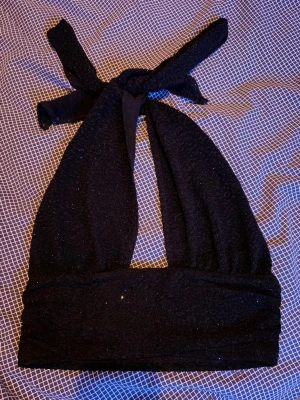 Svart glittrig halterneck topp - Snygg svart halterneck topp med glittereffekt och bred knytning i nacken. Toppen har en croppad passform och är gjord i ett stretchigt, texturerat material. Perfekt för att sticka ut på tjejkvällen eller klubben.