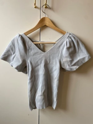 ribbad topp med puffärm - Trendig ribbad topp med v-ringning och puffiga korta ärmar. Toppen har en snygg knytning i ryggen och är tillverkad i ett mjukt, stretchigt material. Perfekt för dig som gillar stilrena detaljer och en feminin siluett. använd ett antal ggr men forf bra skick!