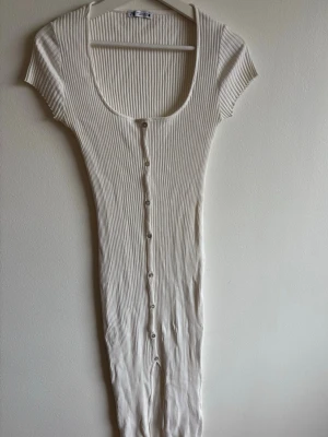 Vit ribbad långklänning med knappar - Snygg vit ribbad Zara långklänning med korta ärmar och djup urringning. Klänningen har knappar framtill hela vägen ner och är figurnära i modellen. Perfekt för dig som gillar en enkel men trendig stil. Knappt använd, lite genomskinlig. Går till knäna.