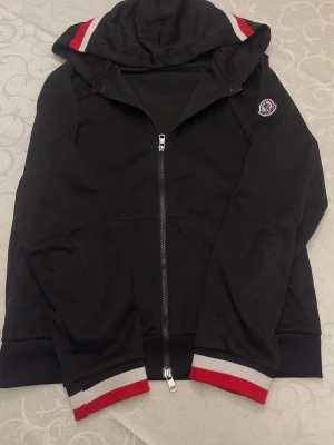 Svart Moncler hoodie med dragkedja - Svart hoodie från Moncler med dragkedja framtill och huva. Ribbade muddar och fåll med vita och röda ränder. Moncler-logga på vänster ärm. Tillverkad i mjukt bomullsmaterial som känns skönt mot huden. Perfekt för en avslappnad och snygg streetwear-look.