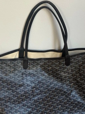 Svart shoppingväska  - Stilren shoppingväska  i svart med klassiskt mönster och vita detaljer. Väskan har dubbla handtag i svart läder och en rymlig form. Perfekt för dig som vill ha en ikonisk och trendig accessoar till vardagen.