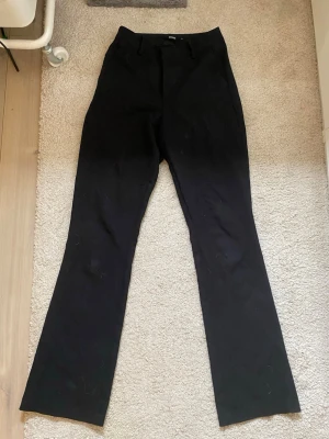 Svarta kostymbyxor - Säljer ett par klassiska svarta kostymbyxor med utsvängda ben och diskreta hällor för bälte. Byxorna har en enkel design och är perfekta för dig som gillar stilrena plagg.