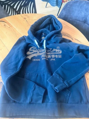 Blå Superdry hoodie med strasslogga - Snygg blå hoodie från Superdry med stor logga i strass på bröstet och japansk text. Klassisk känguruficka framtill, ribbade muddar och dragsko i huvan. Perfekt för en chill och trendig look. Mjuk bomullsblandning som känns skön mot huden.