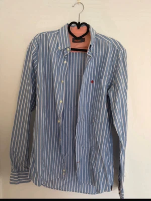 Blåvit randig skjorta från Morris - Snygg blå och vit randig skjorta från Morris med klassisk krage och knappar framtill. Skjortan har lång ärm och en liten röd broderad detalj på bröstfickan. Tillverkad i mjuk bomull, perfekt för en stilren look.