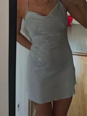 Glittrig silvrig kort klänning - Supersnygg kort klänning med tunna axelband och glittrigt tyg. Klänningen har  öppen rygg med knytband. Storlek xs. Finns en liten reva som knappt synt nu då jag klippt bort den, men ville ändå visa! :) 