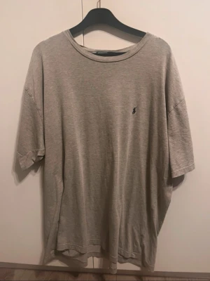 Grå oversized t-shirt från Ralph Lauren - Säljer en grå oversized t-shirt från Ralph Lauren med klassisk rund hals och diskret logga broderad på bröstet. T-shirten är kortärmad och gjord i mjuk bomull, perfekt för en avslappnad och stilren look.