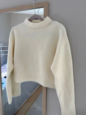 Krämvit croppad stickad tröja - Säljer en krämvit croppad stickad tröja med hög hals och bred ribbstickad kant nedtill samt vid ärmsluten. Tröjan har en mjuk och fluffig känsla och slits i sidorna för en snygg detalj. Perfekt för lager på lager och enkel att styla. Aldrig använd.