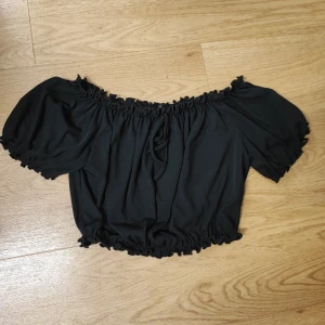 Svart off shoulder crop top - Trendig svart off shoulder crop top med puffärmar och resår vid axlar, ärmslut och midja. Toppen har en liten rosett framtill och är gjord i ett mjukt, lätt tyg som ger en avslappnad men snygg look. 