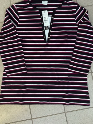 Randig långärmad topp från et! - Säljer en långärmad topp från et! i svart, vit och rosa med horisontella ränder. Toppen har v-ringning med dekorativa metallknappar längs öppningen. Tillverkad i mjuk bomull, perfekt för en avslappnad och trendig look.