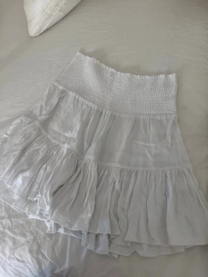 Vit volangkjol med smock - Säljer en superfin vit kortkjol med bred smock i midjan och flera lager volanger. Kjolen har en luftig och flowy känsla, perfekt för varma dagar. Materialet känns lätt och bekvämt, och den passar perfekt till en avslappnad sommarstil.