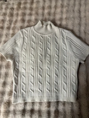 Vit kabelstickad topp med kort ärm - Kabelstickad topp i vitt med korta ärmar och hög ribbad krage. Tillverkad i en mjuk blandning av viskos, polyester och nylon. Snygg struktur med klassiskt mönster framtill och ribbade detaljer vid ärmslut och nederkant. Storlek S men ser ut som en XXS tröja på och utan.