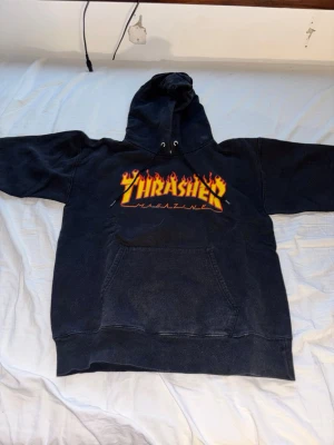 Svart Thrasher hoodie  - Svart hoodie från Thrasher med den ikoniska loggan i gult och rött flames-tryck på bröstet. Klassisk känguruficka framtill och justerbar huva med snören. Perfekt för dig som gillar streetwear och skatekultur.