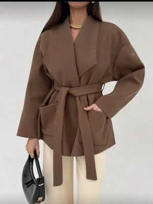 Brun omlottkappa med knytskärp - Snygg brun kappa i omlottmodell med brett slag och knytskärp i midjan. Kappan har två stora framfickor och är tillverkad i ett mjukt tyg som ger en avslappnad men stilren look. Perfekt för dig som gillar minimalistisk stil.