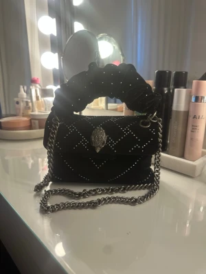 Svart handväska med kedja och glittrande detaljer  - Snygg svart handväska i sammet med silverfärgade nitar i rutmönster. Väskan har en fluffig scrunchie-inspirerad handtag och en lång silverkedja för axeln. Framtill finns ett dekorativt spänne i form av en hand. Perfekt för dig som gillar statement-accessoarer. Enstak av de glittrande detaljerna har gått bort men inget man märker 