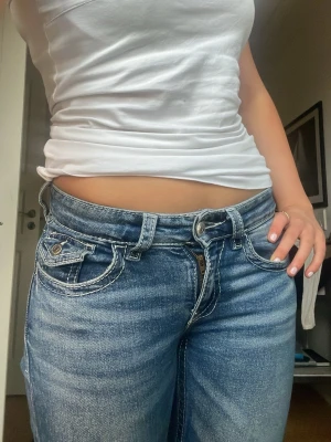 Blå bootcut jeans med kontrastsömmar - Säljer ett par blå bootcut jeans med tydliga kontrastsömmar och fem fickor. Jeansen har låg midja och slitna detaljer för en avslappnad look. Perfekta för dig som gillar en klassisk men trendig stil.