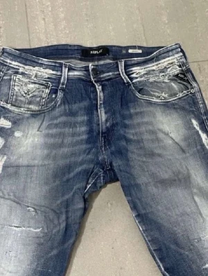 Blå slitna jeans från Replay - Säljer ett par blå jeans från Replay med slitningar och tvättade detaljer för en cool, avslappnad look. Jeansen har fem fickor, normal midja och raka ben. Perfekta för dig som gillar streetwear och en trendig stil.
