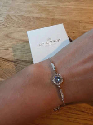 Armband - Armband Lilly and Rose. I färgen silver och blå. Vackert armband som är i färgen silver och en ljusblå mittsten. Saknas dock en sten på ena sidan, se bilden. Syns knappt för det blänker så mycket.