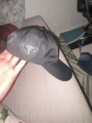 Svart keps från Prada i nylon - Snygg svart keps från Prada med klassisk böjd skärm och ikoniska metalltriangeln på sidan. Tillverkad i slitstarkt nylonmaterial och justerbar baktill för perfekt passform. Perfekt accessoar för en clean och stilren look.