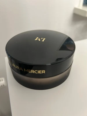 Laura Mercier Translucent Loose Powder - Translucent Loose Setting Powder från Laura Mercier i en rund burk med svart lock och guldfärgad text. Pudret är transparent och ger en naturlig finish. Förpackningen innehåller 49 g och är Anniversary Edition. Perfekt för att fixera sminket och matta ner huden. Oanvänd (plasten är kvar på insidan), nypris 829kr