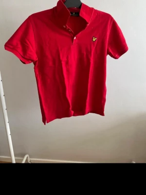 Röd kortärmad skjorta från Lyle & Scott - Snygg röd skjorta från Lyle & Scott med klassisk krage och korta ärmar. Skjortan har knappar framtill och den ikoniska fågel-loggan på bröstet. Perfekt för dig som vill ha en enkel och stilren look.