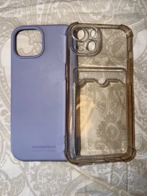Två mobilskal, lila & transparent - Säljer två mobilskal till iPhone. Ett är lila i mjukt silikon från MobileFriend och det andra är transparent med kanter i hårdplast, har även ett kortfack på baksidan. Tillhör iPhone 13 