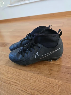 Svarta Nike fotbollsskor med snörning - Säljer ett par svarta Nike fotbollsskor med klassisk vit swoosh på sidan. Skorna har snörning, högre strumpliknande skaft och dobbar för bra grepp på planen. Tillverkade i syntetmaterial och designade för att ge stabilitet och kontroll under matchen. Str 36.5