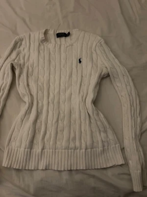 Vit kabelstickad tröja Polo Ralph Lauren - Kabelstickad vit tröja från Polo Ralph Lauren med rund halsringning och lång ärm. Tröjan har det klassiska broderade logotypen på bröstet och ribbstickade muddar vid ärmslut och nederkant. 