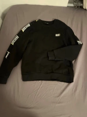 Svart sweatshirt från EA7 Emporio Armani - Svart sweatshirt från EA7 Emporio Armani med logga på bröstet och tryck längs båda ärmarna. Rund halsringning och ribbade muddar vid ärmslut och nederkant. Perfekt för en sportig och avslappnad stil.