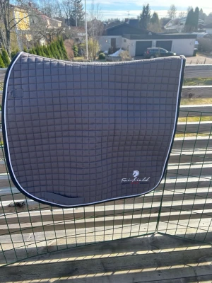 Grått schabrak från FairField - Säljer ett stilrent grått schabrak från FairField med rutmönster och vit kant. Perfekt till hästutrustning och ger en snygg look till sadeln. Materialet är quiltat tyg och schabraket har en diskret logga i hörnet. Kardborrebanden för att fästa schabraket är avklippta men det sitter ändå väldigt bra! 