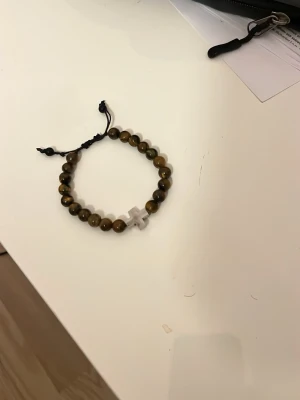 Brunt pärlarmband med kors - Snyggt armband med bruna pärlor och ett kors i mitten. Pärlorna har en glansig finish och armbandet har justerbart svart snöre. Perfekt accessoar för dig som gillar en stilren och personlig look.