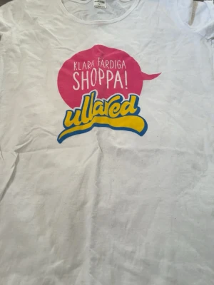 Vit Ullared t-shirt med tryck - Vit t-shirt från Ullared med färgglatt tryck framtill där det står 'KLARA FÄRDIGA SHOPPA!' i rosa och 'ullared' i gul och blå text. Klassisk passform och korta ärmar. Perfekt för dig som gillar shopping och vill ha en rolig statement-tee.