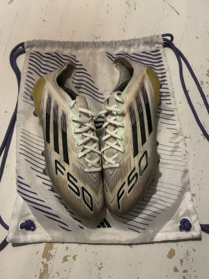 Adidas F50 fotbollsskor vita/guld elite - Säljer ett par Adidas F50 fotbollsskor i vitt med svarta detaljer och guldfärgad häl. Skorna har klassiska svarta stripes och texten F50 på ovansidan. Medföljer gör en matchande vit påse och lila innersulor. Perfekta för dig som vill sticka ut på planen.