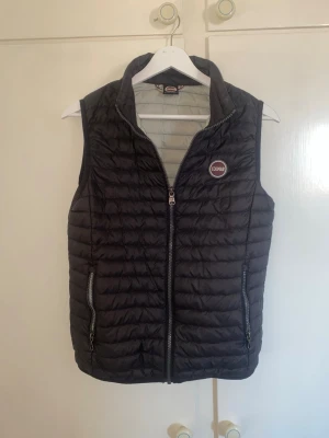 Svart dunväst från Colmar - Snygg svart dunväst från Colmar med quiltad design och två dragkedjeförsedda fickor framtill. Västen har hög krage och Colmar-logga på bröstet. Perfekt att slänga över en hoodie eller tröja för extra värme. Materialet är lätt och smidigt. Storlek 166 cm/xs