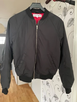 Svart bomberjacka från Stay - Snygg svart bomberjacka från Stay med klassisk siluett, ribbade muddar och dragkedja framtill. Jackan har två snedställda fickor och röd innerfoder som ger en cool kontrast. Tillverkad i ett slätt, tåligt material som passar perfekt till streetwear-stilen.