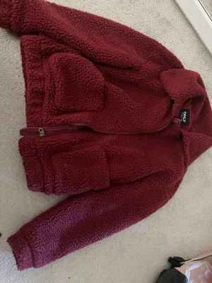 Vinröd teddyjacka från ONLY - Mysig vinröd teddyjacka från ONLY med dragkedja framtill och stora fickor. Jackan har en hög krage och är gjord i fluffigt teddy-material som håller dig varm. Perfekt för dig som vill ha en trendig och bekväm look under kyliga dagar.pris kan såklart diskuteras 
