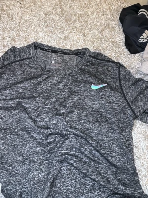 Gråmelerad Nike Dri-Fit t-shirt - Snygg gråmelerad t-shirt från Nike med Dri-Fit-teknologi. T-shirten har korta ärmar, rund halsringning och en liten Nike-logga i turkos och rosa på bröstet. Perfekt för träning eller chill dagar.