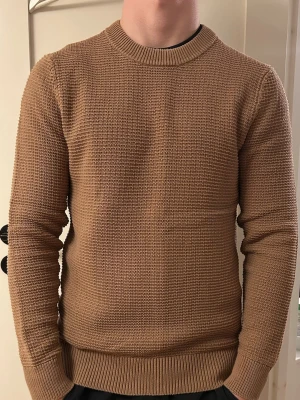 Beige stickad tröja J.Lindeberg - Säljer en ljusare brun/mörk beige stickad tröja från J.Lindeberg. Tröjan har rund halsringning, ribbade muddar och nederkant samt en grovstickad struktur. Perfekt för dig som gillar stilrena och klassiska plagg i garderoben.