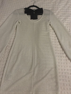 Glittrig vit långärmad klänning - Säljer en glittrig vit kortklänning med lång ärm. Klänningen har en elegant svart rosett i ryggen och är perfekt för dig som vill sticka ut. Materialet känns mjukt och skimrar snyggt i ljuset.