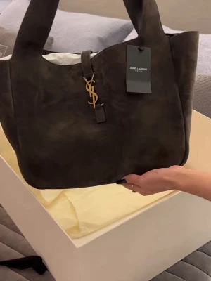 Mörkbrun Saint Laurent handväska - Säljer en snygg mörkbrun handväska från Saint Laurent i mjukt mockamaterial. Väskan har en stilren, rymlig design med breda axelband och ikoniskt YSL-guldemblem framtill. Perfekt för dig som vill ha en lyxig och trendig accessoar.