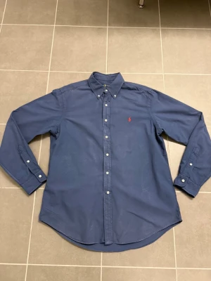Blå skjorta från Ralph Lauren, stl L - Klassisk blå skjorta från Ralph Lauren med button-down krage och vit knappstängning. Skjortan har lång ärm och den ikoniska röda logotypen broderad på bröstet. Tillverkad i bomull för en bekväm och stilren look.