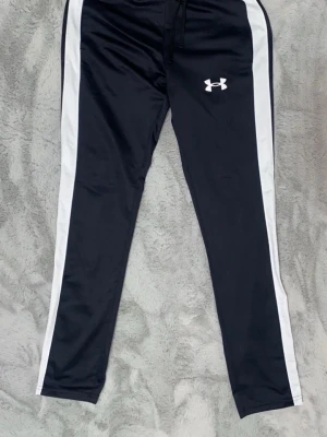 Svarta träningsbyxor Under Armour - Snygga svarta träningsbyxor från Under Armour med vita sidopaneler och logga på vänster lår. Byxorna har smal passform, resår i midjan och dragsko. Tillverkade i ett mjukt och stretchigt material som passar perfekt till träning eller sport. Helt nya!!!