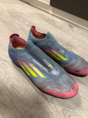 Adidas F50 blå/rosa fotbollsskor ag elite - Säljer ett par Adidas F50 fotbollsskor i blått och rosa med gula detaljer. Skorna har en strömlinjeformad design utan snörning och är tillverkade i ett lätt syntetmaterial för maxad speed på planen. Perfekta för dig som vill sticka ut och spela snabbt.