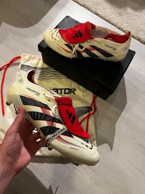 Adidas Predator fotbollsskor  - Säljer ett par Adidas Predator fotbollsskor i vitt med svarta och röda detaljer. Skorna har klassisk Predator-design med röd plös, svarta ränder och texten Predator på sulan. Tillverkade i syntetmaterial och utrustade med fasta dobbar för gräsplan.